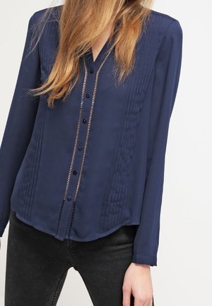 Blusa a maniche lunghe di colore blu navy con chiusura a bottoni, dettagli a pieghe e texture liscia. Pannelli trasparenti sottili e orlo arrotondato aggiungono particolari.