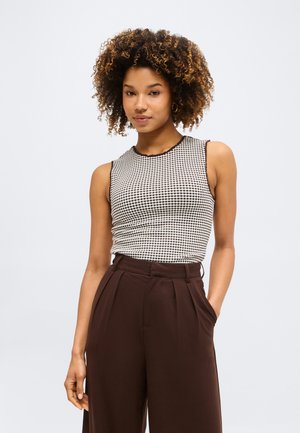 Γυναίκα με σγουρά μαλλιά που φοράει ένα αμάνικο ασπρόμαυρο ριγέ τοπ και καφέ παντελόνι με φαρδιά πόδια και ψηλή μέση, στέκεται με το ένα χέρι στην τσέπη.