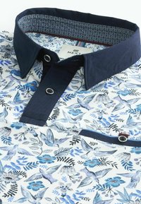 Chemise homme avec col bleu marine et patte de boutonnage, tissu blanc imprimé d’oiseaux bleus et gris ainsi que de fleurs, avec un petit détail de poche boutonnée.