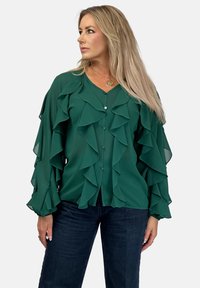 Zielona bluzka z falbanami, wykonana z lekkiego materiału, z zapięciem na guziki. Posiada długie, rozszerzane rękawy oraz dekolt w kształcie litery V. Połączona z ciemnymi dżinsami.