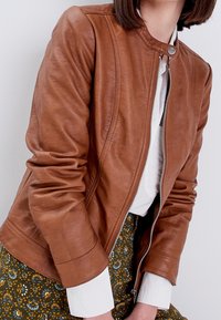 Veste en cuir marron avec un col rond, une fermeture éclair à l'avant et des manches froncées, portée sur une jupe à motifs avec des détails floraux.