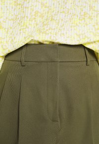 Chemisier à motifs jaunes et blancs rentré dans un pantalon taille haute vert olive avec passants de ceinture et plis avant.