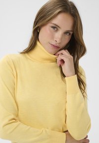 Pull à col roulé jaune en tissu tricoté doux ; col et poignets côtelés ; coupe ajustée avec une texture lisse et des variations subtiles.