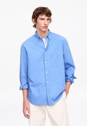 Jeune homme portant une chemise bleue ample boutonnée sur un maillot blanc, manches retroussées et pantalon clair sur fond blanc.