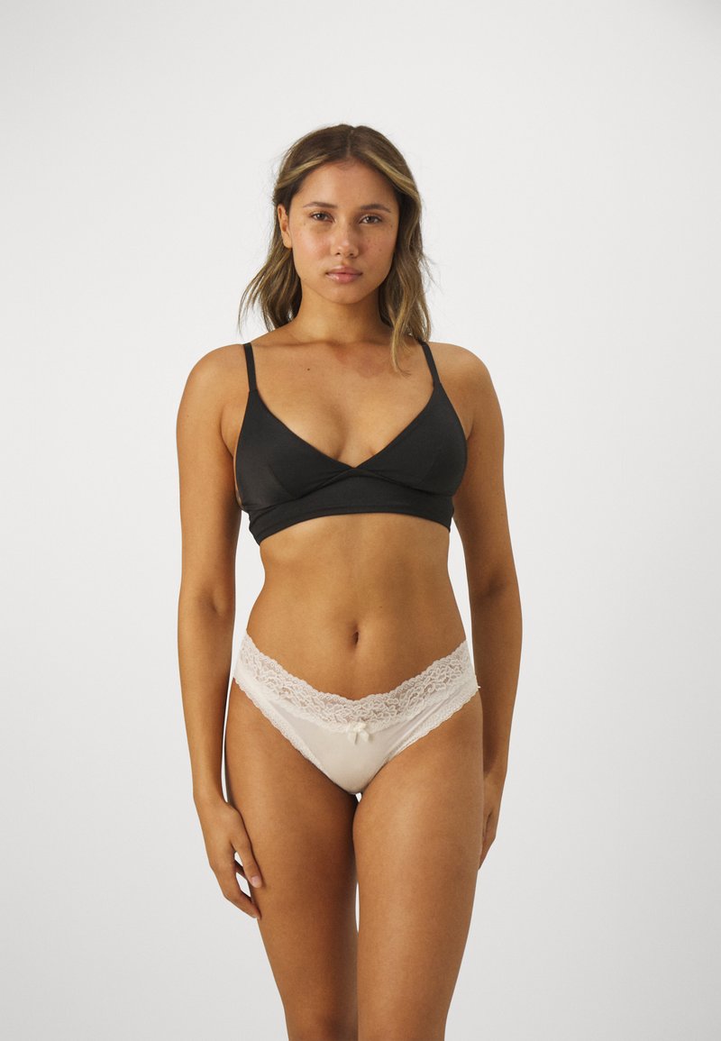 Svart korsremad bralette i mjukt tyg. Cremefärgad stringtrosa med spetskant och rosettdetalj i centrum. Slät textur, minimalistisk design.