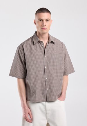 Jonge man met kort haar, gekleed in een losse bruine geruite korte mouwen overhemd en witte broek, staand tegen een effen achtergrond.