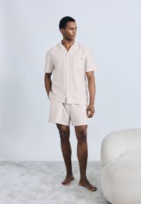 Φωτεινό μπεζ κοντομάνικο πουκάμισο και ταιριαστό σορτς με λεπτές κατακόρυφες ρίγες, κατασκευασμένα από απαλό ύφασμα, με τσέπη στο στήθος και διακριτικό λογότυπο.