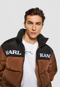 Ruda ir juoda pūkinė striukė su užtrauktuku, ant krūtinės su balta siuvinėta inscription "KARL", dėvima ant baltos marškinėlių.