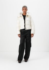 Witte pufferjack met een capuchon, voorzien van een rits aan de voorkant en twee voorkruiden. Draagt een zwarte top en is gecombineerd met losse zwarte broek.