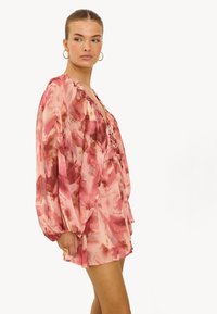 Vrouw in een losse, lange mouwen, roze en rode bloemenpatroon korte jurk met een gerimpelde halslijn, gouden hoepelringen en een gouden ring.