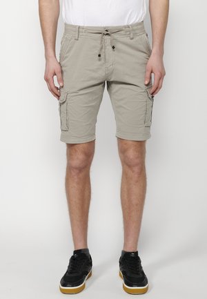 Homme portant un short cargo beige avec taille à cordon et des baskets noires avec semelles en caoutchouc, se tenant contre un fond blanc.