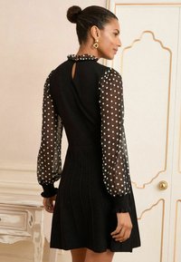 Black dress with a fitted bodice and flared skirt, featuring sheer polka dot sleeves, ruffled neckline, and back keyhole detail.