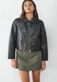 Giacca in pelle nera con colletto, design corto e zip frontale. Indossata sopra un vestito verde texturizzato con righe verticali.