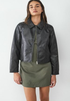 Schwarze Lederjacke mit kragen, taillierter Form und Reißverschluss vorne. Getragen über einem strukturierten grünen Kleid mit vertikalen Streifen.