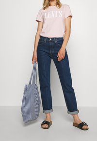 Pink t-shirt with white "LEV'S" logo, dark blue high-waisted jeans, black slip-on sandals, and a striped blue and white tote bag.
