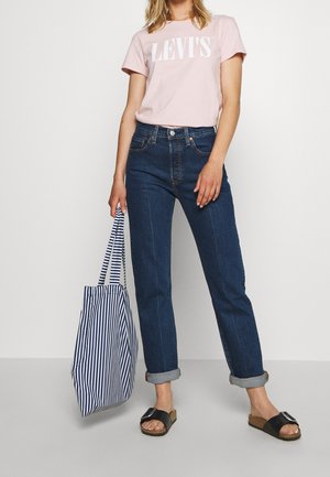 Pink t-shirt with white "LEV'S" logo, dark blue high-waisted jeans, black slip-on sandals, and a striped blue and white tote bag.