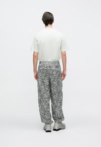 Egy férfi hátulról áll, egyszerű világos színű pólót, fekete-fehér mintás nadrágot és világosszürke sportcipőt visel.