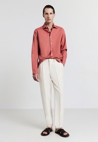 Giovane con capelli tirati indietro, indossa una camicia button-up color ruggine, pantaloni plissettati crema e sandali intrecciati neri, in piedi davanti a uno sfondo semplice.