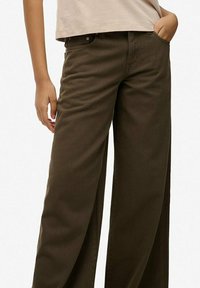 Persoon draagt een donkerbruine broek met wijde pijpen en een beige cropped top, met één hand in de zak en de andere ontspannen langs het lichaam.