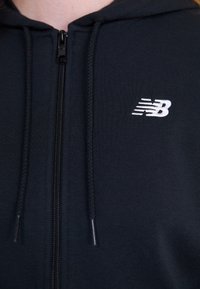 Svart zip-up hettegenser laget av mykt stoff, med en liten hvit New Balance-logo på venstre bryst og snorhette.