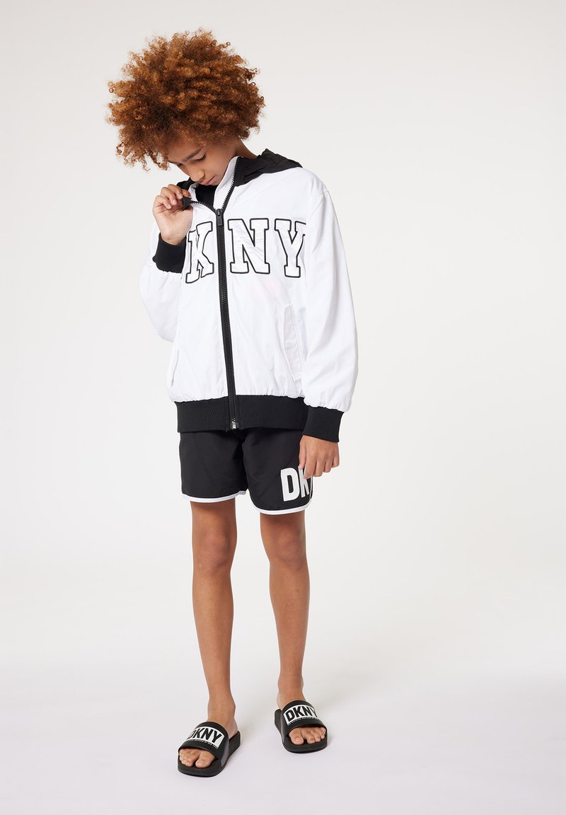 Giacca bomber bianca con zip, dettagli neri e grande logo "DKNY"; abbinata a pantaloni corti neri con logo e bordi bianchi, e ciabatte nere.