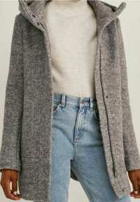 Manteau gris en mélange de laine avec une surface texturée et duveteuse, une fermeture éclair, des poches latérales et une capuche, superposé sur un pull en tricot crème et un denim clair.