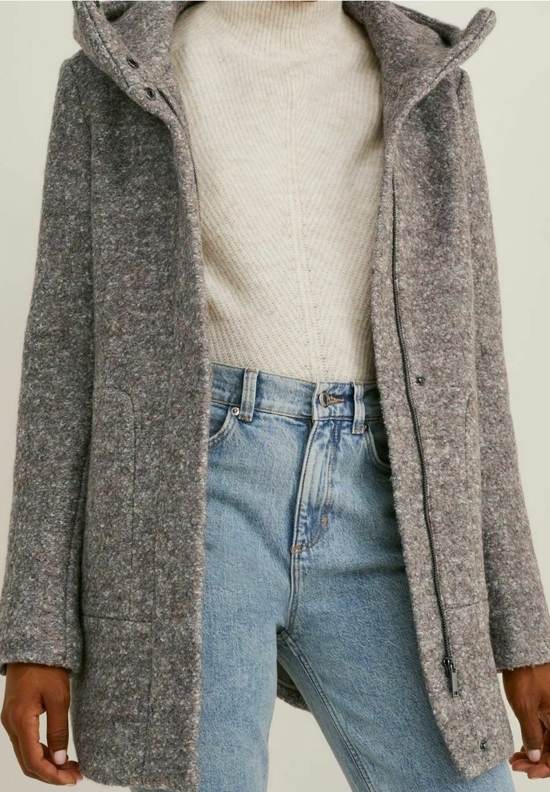 Manteau gris en mélange de laine avec une surface texturée et duveteuse, une fermeture éclair, des poches latérales et une capuche, superposé sur un pull en tricot crème et un denim clair.