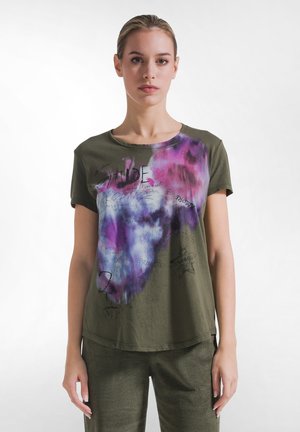 Mujer con cabello claro que lleva una camiseta verde oliva con patrones abstractos en púrpura, rosa y blanco y texto manuscrito negro en el frente.