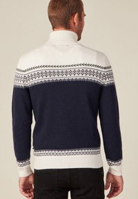 Pull en tricot avec col montant, corps bleu marine foncé, manches blanches avec bande à motifs, et ourlet inférieur en côte contrasté.