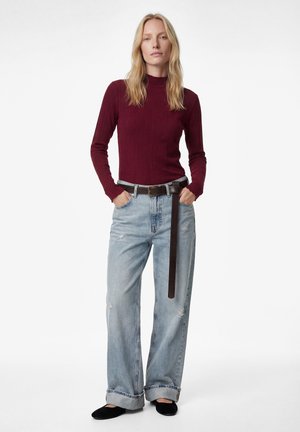 Kvinde med langt blond hår iført en vinrød ribstrikket sweater, lyseblå vidde jeans med et langt mørkt bælte og sorte sko, stående med hænderne i lommerne.