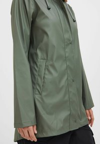 Green waterproof jacket made of shiny material, featuring snap buttons, a hood with cords, and a side pocket on the left.