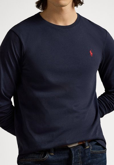 Tengerészkék hosszú ujjú póló puha pamutból, kerek nyakkivágással és a mellkason egy kis piros hímzett logóval.