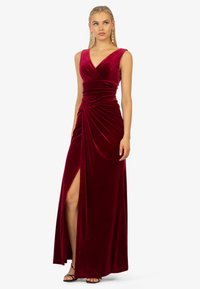 Bordeaux rotes Samt-Maxikleid mit V-Ausschnitt, taillierter Taille, drapierten Details und einem seitlichen Schlitz bis zur Oberschenkelhöhe. Schwarze Riemchen-Heels runden den Look ab.
