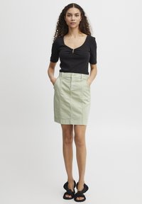 Haut noir ajusté avec un détail en trou de serrure, associé à une jupe longueur genoux vert clair avec des poches avant et une fermeture à boutons. Sandales noires.