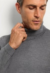Grauer Rollkragenpullover mit gerippter Textur, im figurbetonten Design und mit hohem Kragen. Modell greift sanft mit einer Hand am Kragen.