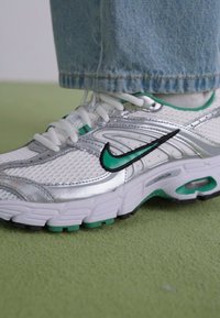 White athletic shoe with silver accents, green details, mesh upper, and cushioned sole. Notable swoosh logo and textured surface.