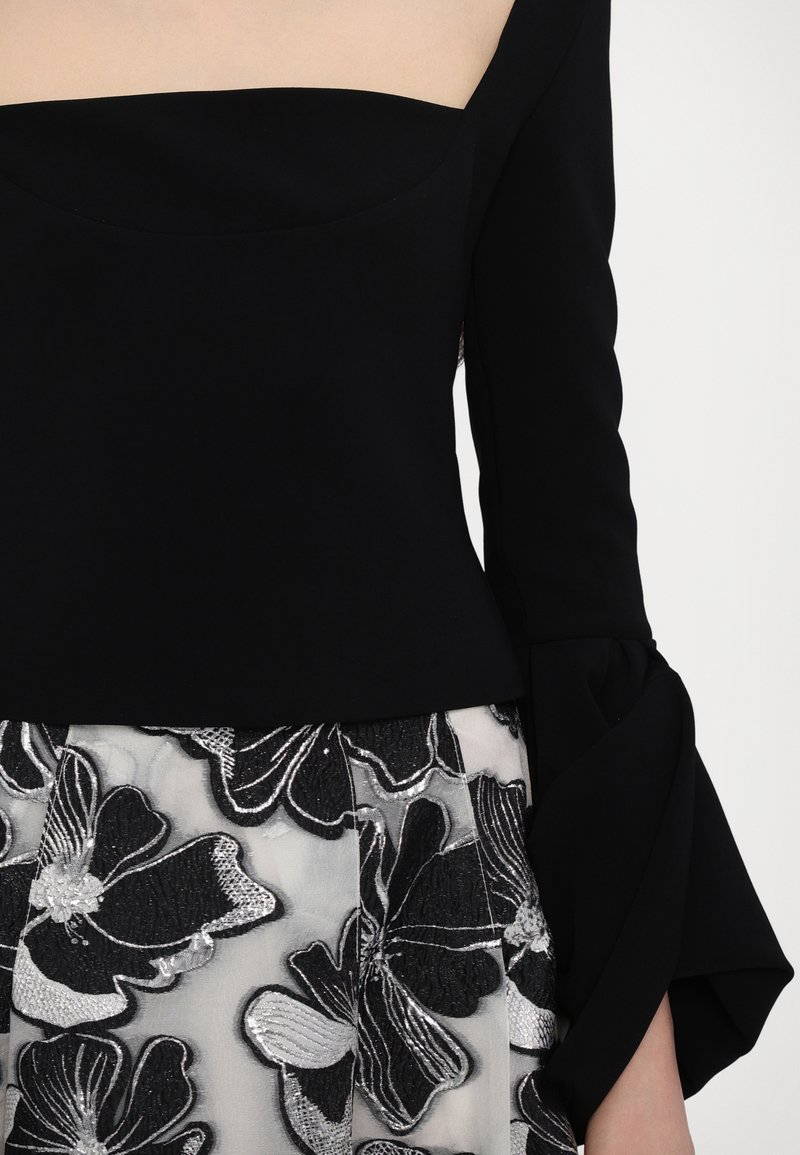 Top nero con scollatura quadrata e maniche lunghe, abbinato a una gonna trasparente con motivi floreali ricamati in argento.