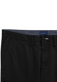 Černé kalhoty s hladkým povrchem, s denimovým páskem, předními kapsami a zapínáním na knoflík, na kterém je viditelná značka.