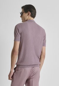 Hombre que lleva un suéter lila de manga corta con textura acanalada y pantalones a juego, de espaldas, con gafas de sol oscuras y el cabello bien peinado.