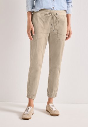 Person iført beige cargobukser med justerbar snor og elastiske manchetter samt beige snøresneakers, stående mod en ensfarvet baggrund.