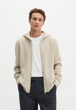 Jonge man die een beige zip-up hoodie draagt over een wit overhemd en donkere broek, staand met handen in de zakken tegen een effen achtergrond.