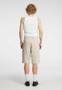 Beżowe spodenki do kolan wykonane z teksturowanego materiału, z dwoma tylnymi kieszeniami, w zestawieniu z gładkim białym topem na ramiączkach i czarnymi butami.