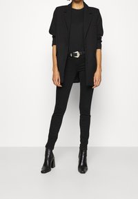 Blazer negro sobre una blusa negra, combinado con jeans ajustados negros y botas de tacón tobilleras. Cinturón metálico añade un toque de detalle.