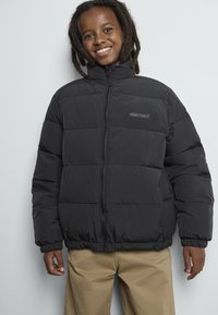 Smilende barn med skulderlange dreadlocks iført en svart boblejakke og beige bukser mot en lys bakgrunn.