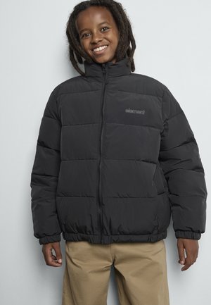 Smiling child with shoulder-length dreadlocks wearing a black puffer jacket and beige pants against a plain light background.