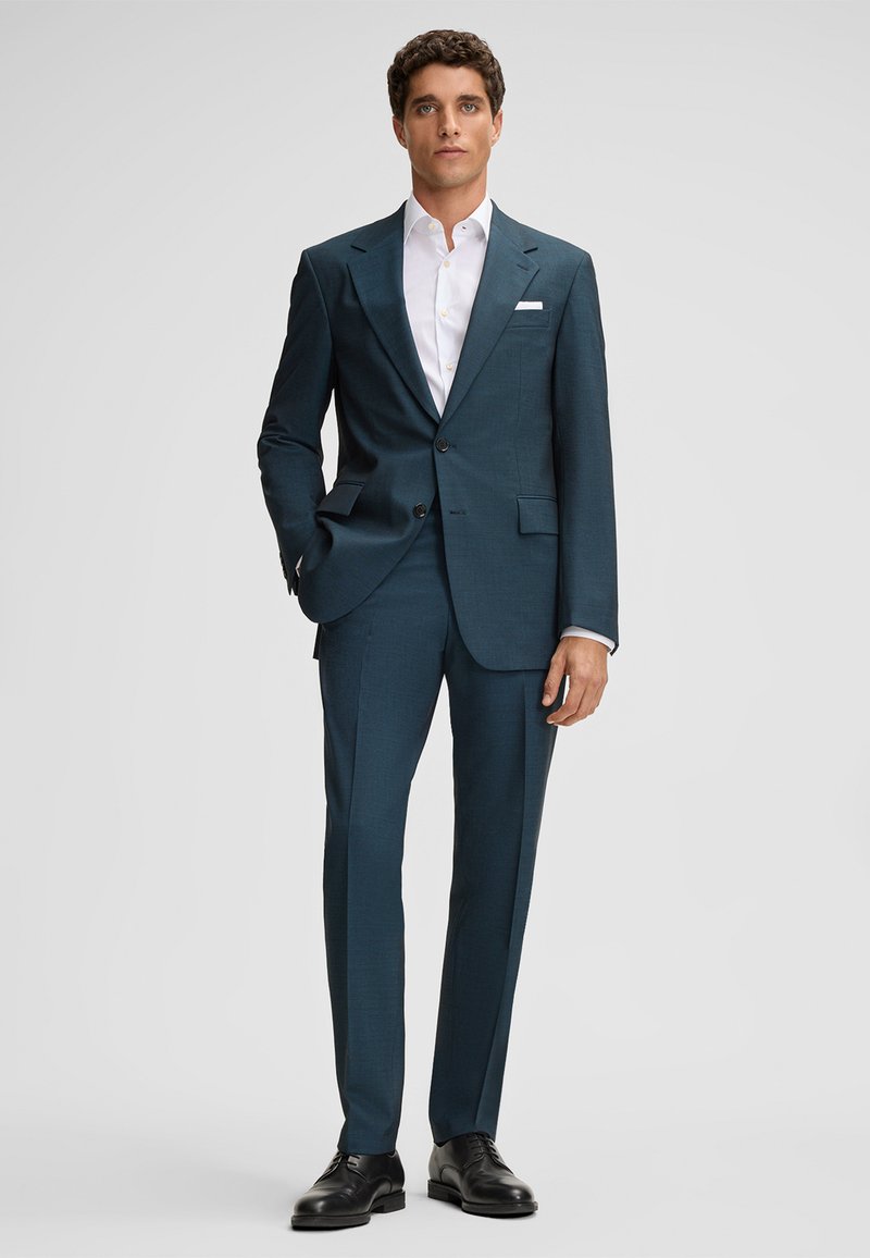 Man staat in een donkerblauw groen pak met een wit shirt, zwarte dresschoenen en een witte pochet op een effen lichte achtergrond.