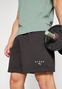Shorts de sport noirs avec le texte et le logo "HYROX" à l'avant, fabriqués en matériau texturé, associés à un t-shirt gris et à un poids.