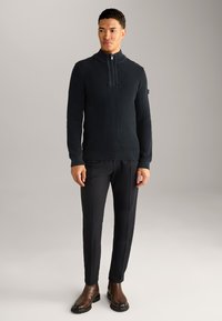 Dunkelblauer gerippter Pullover mit hohem Kragen und Reißverschluss, kombiniert mit schwarzen maßgeschneiderten Hosen und braunen Stiefeletten. Sanfte Textur, taillierte Passform.