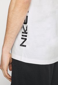 Bílý bavlněný tričko s černým logem Nike potisknutým vertikálně na boku. Nosené s černými síťovanými šortkami, ukazujícími jednoduchý design.