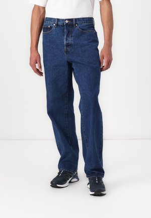 Homme portant un jean droit bleu foncé avec des baskets noires et blanches, debout devant un fond blanc uni.